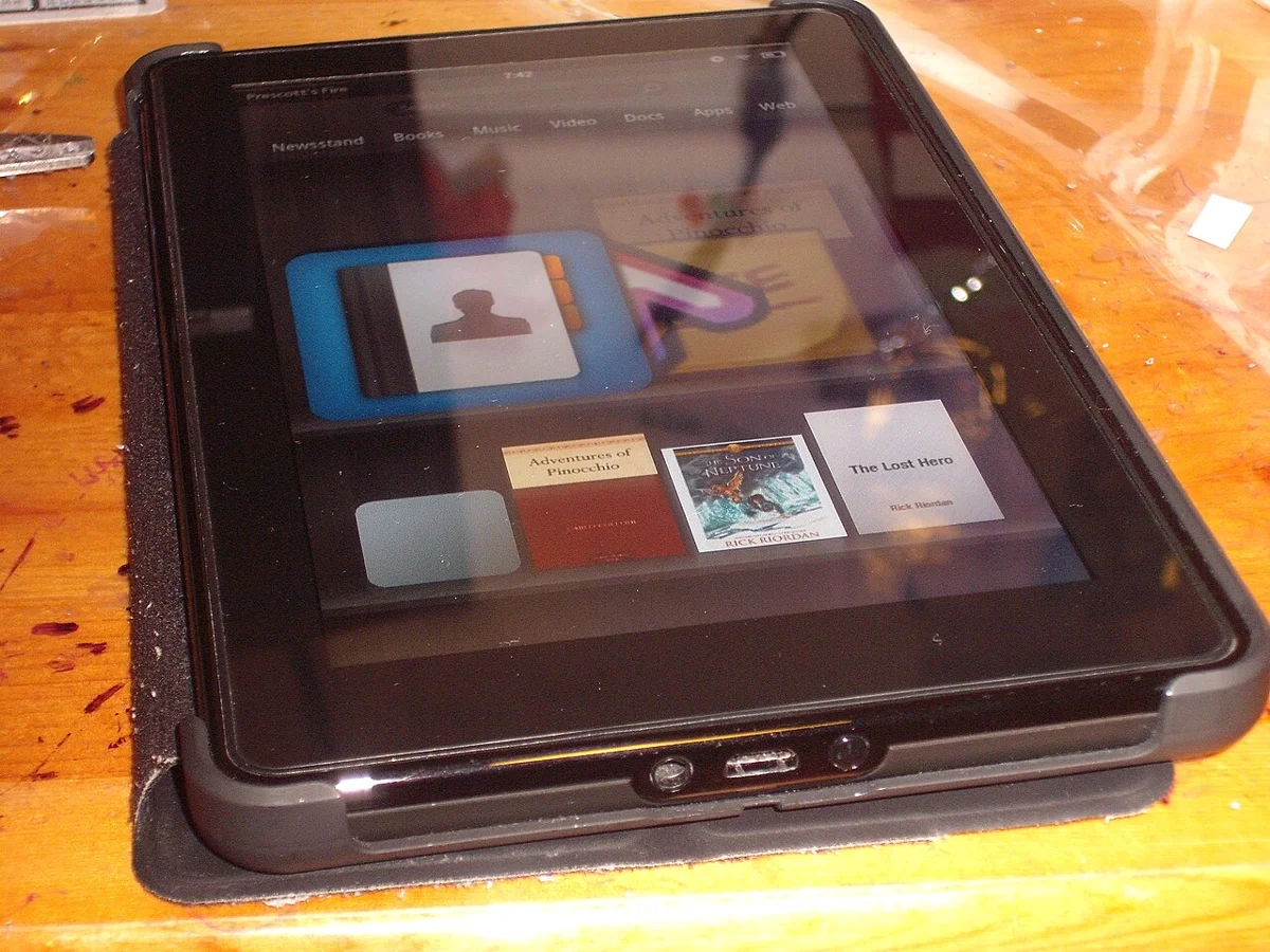 A real photo of an older Amazon Kindle Fire tablet with its cover attached, placed on a table, showing a book app and books on the screen. This is a reference photo of an older Fire device, not the new model.