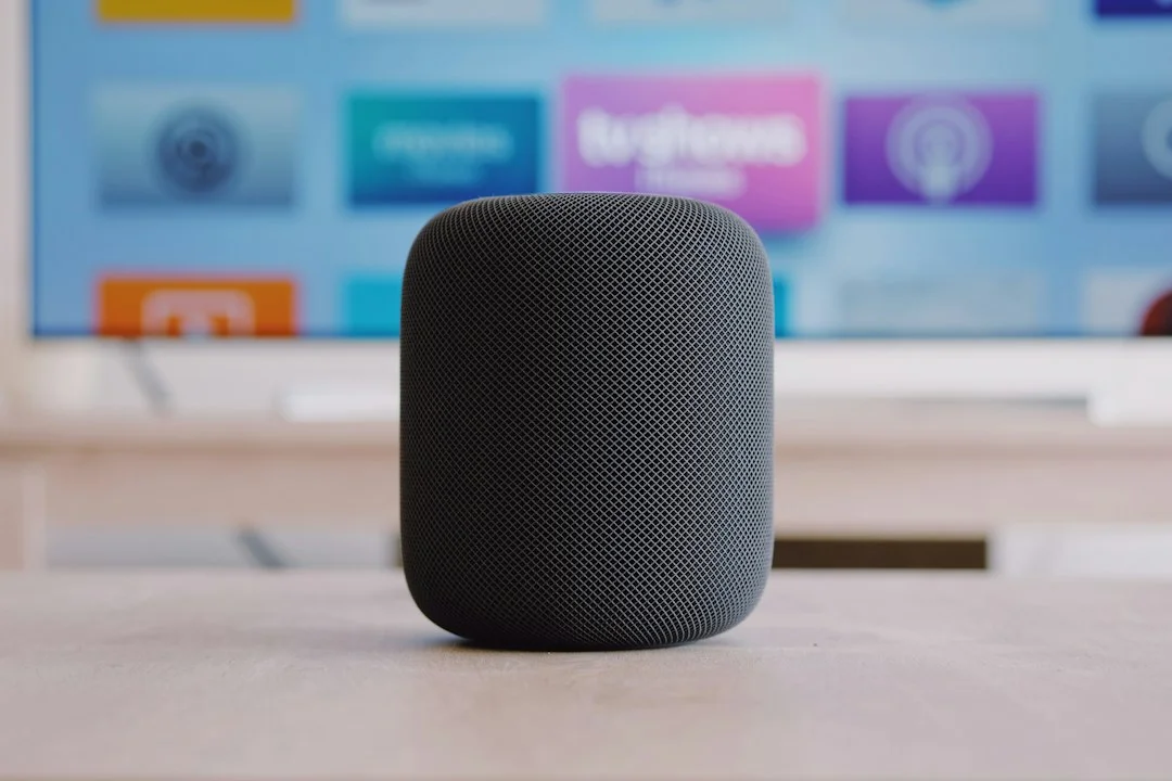 A single black Apple HomePod speaker placed on a table in front of a TV showing the Apple TV screen. The number and arrangement in the photo do not represent a four-speaker surround configuration; for reference only.