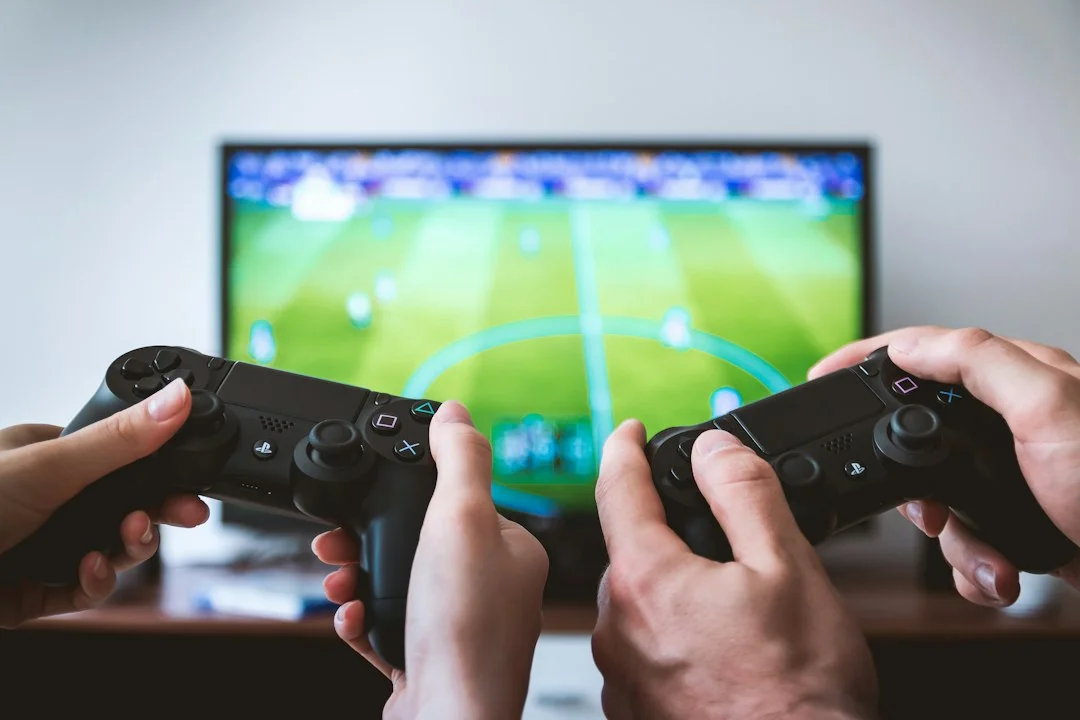 Two hands holding wireless controllers in front of a TV showing a blurred soccer game. A general gaming scene used as a neutral reference photo; it does not show GTA 6 or the leaked video.