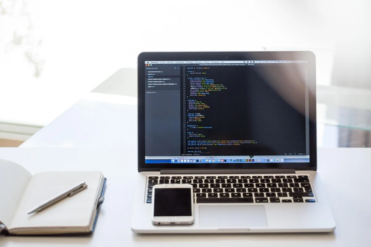 A bright work desk with a laptop displaying code, a notebook, and a smartphone. A neutral reference photo for the section covering game development progress; it does not show Eric Barone himself or an actual development site.