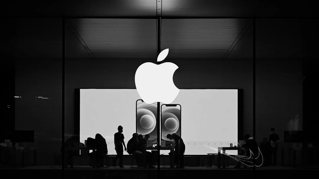 A black-and-white photo of a store glass window bearing the Apple logo, with a large screen and smartphone images displayed inside and silhouettes of people visible. It is not a photo of an actual announcement stage and is a neutral reference photo unrelated to the rumored Apple event on October 27.