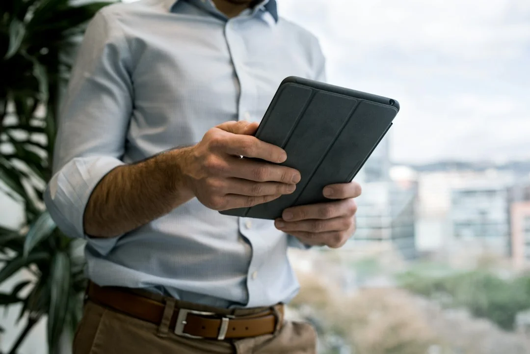 A person holding a small tablet with both hands, wearing a light shirt, standing indoors near a window with a blurred city view. A generic photo illustrating small-tablet use; it does not show the specific model or buyer discussed in the article.
